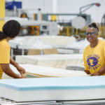 Two workers smile brightly while holding a large piece of cloth at a manufacturing facility in the Central SC Region
